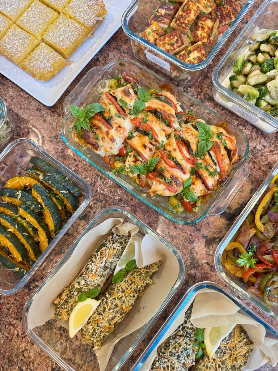 A variety of gourmet prepared dishes including tomato basil chicken, pesto salmon, baked acorn squash, and lemon bars in rectangular glass containers.