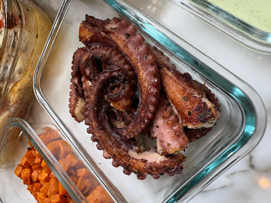 Glass meal prep containers filled with golden-brown Portuguese garlic seared octopus tentacles and sides of vibrant roasted cubed sweet potatoes resting on marble.