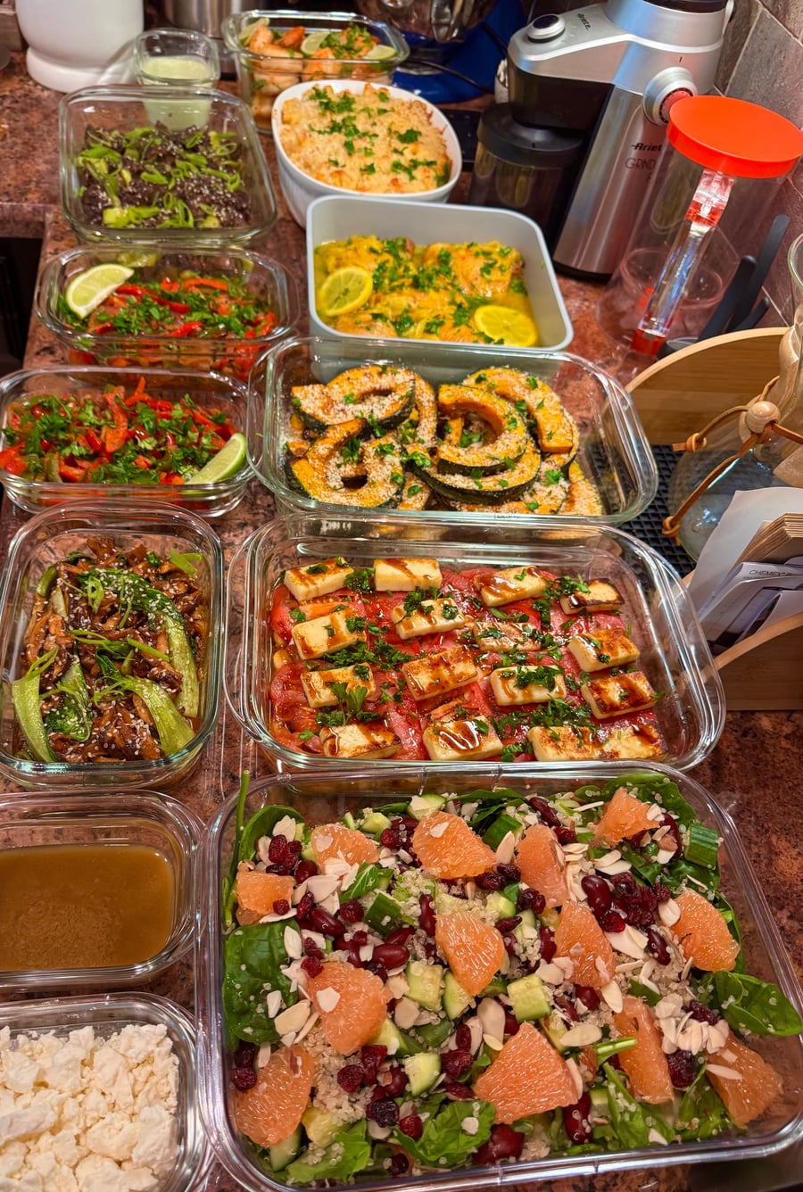 A variety of glass containers filled with roasted squash, grilled halloumi salad, citrus spinach salad, stir-fried beef, and chicken, neatly arranged for meal prep.