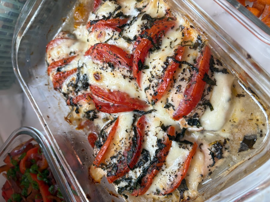 A glass container holding golden baked chicken breasts topped with sliced red tomatoes, melted cheese, and chopped basil leaves, presented for easy serving.