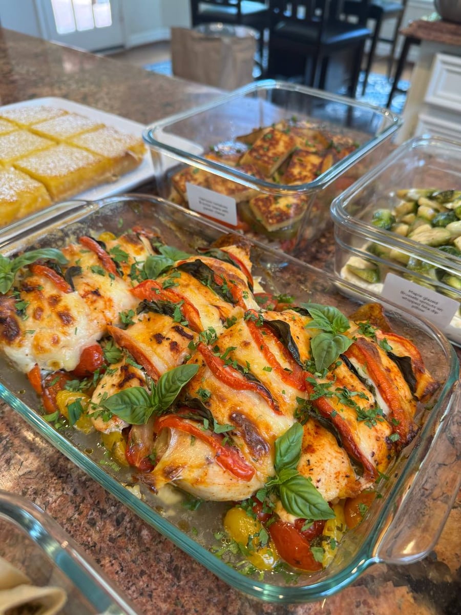 A glass baking dish containing tomato basil chicken breasts topped with melted cheese, fresh basil leaves, and roasted heirloom tomatoes in a professional kitchen setting.