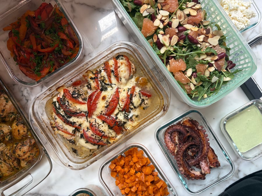 A collection of glass containers featuring tomato basil chicken, garlic seared octopus, roasted sweet potatoes, fajita-style peppers, and a vibrant salad with grapefruit and almonds.