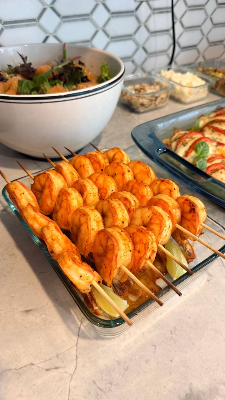 A glass dish filled with paprika-dusted shrimp skewers garnished with fresh lime wedges, alongside a bowl of supergreens salad and tomato basil chicken.