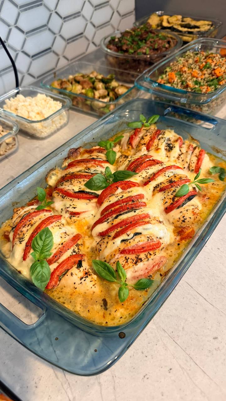 A glass casserole dish features baked tomato basil chicken breasts topped with melted cheese and fresh basil leaves, next to containers of roasted vegetables and quinoa.