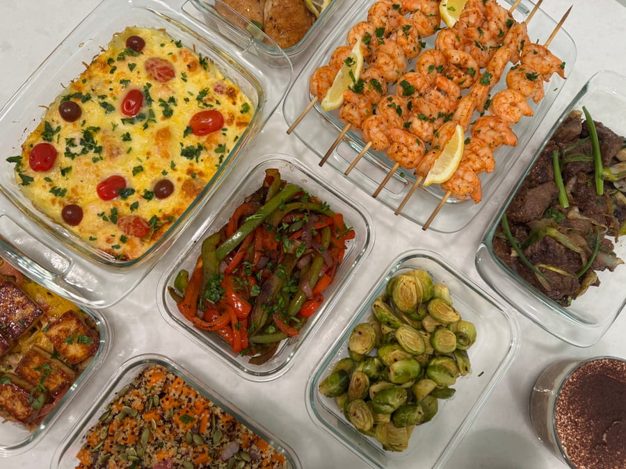 Top-down view of stacked glass containers featuring shrimp skewers, beef stir fry, salmon frittata, roasted sweet potato quinoa, peppers, and brussels sprouts.