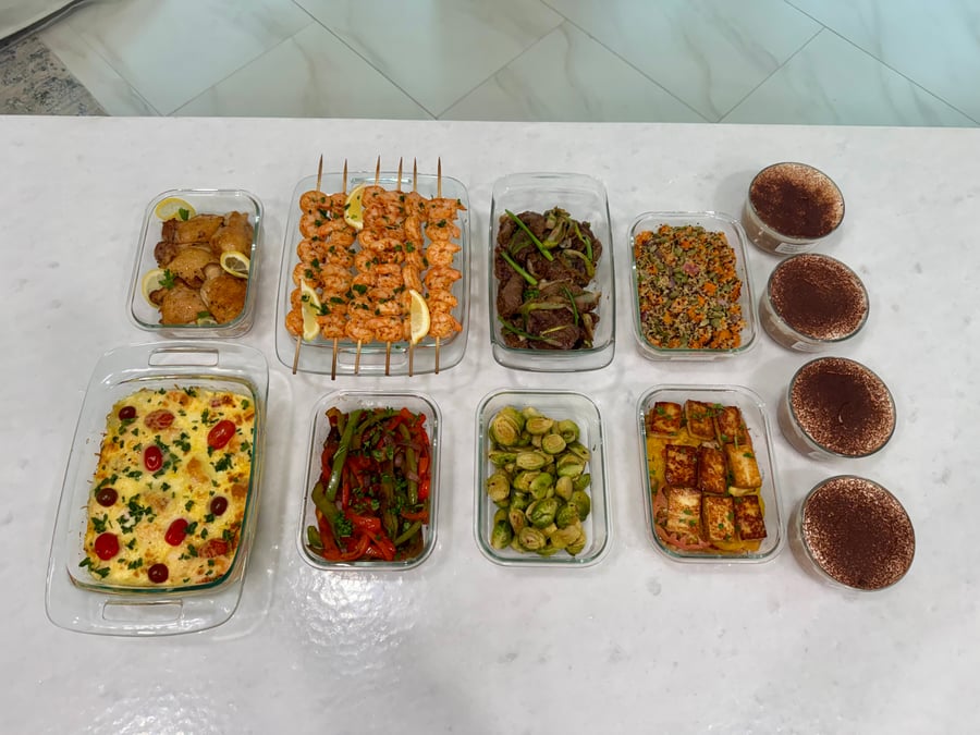 A variety of prepared meals in glass containers featuring shrimp skewers, beef stir fry, salmon frittata, roasted vegetables, and tiramisu protein overnight oats.
