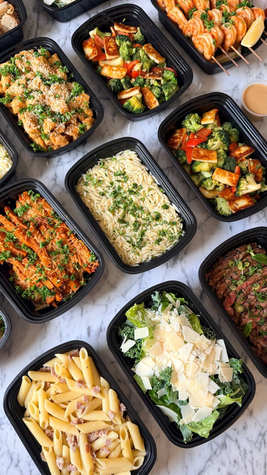 Top-down view of black plastic meal prep containers filled with orange glazed chicken, flank steak, shrimp skewers, pasta, and crisp green salads.