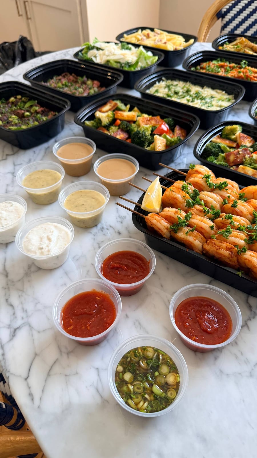 Black containers arranged on a marble surface featuring paprika lime shrimp skewers, sautéed broccoli with paneer, and various house-made dipping sauces and dressings.