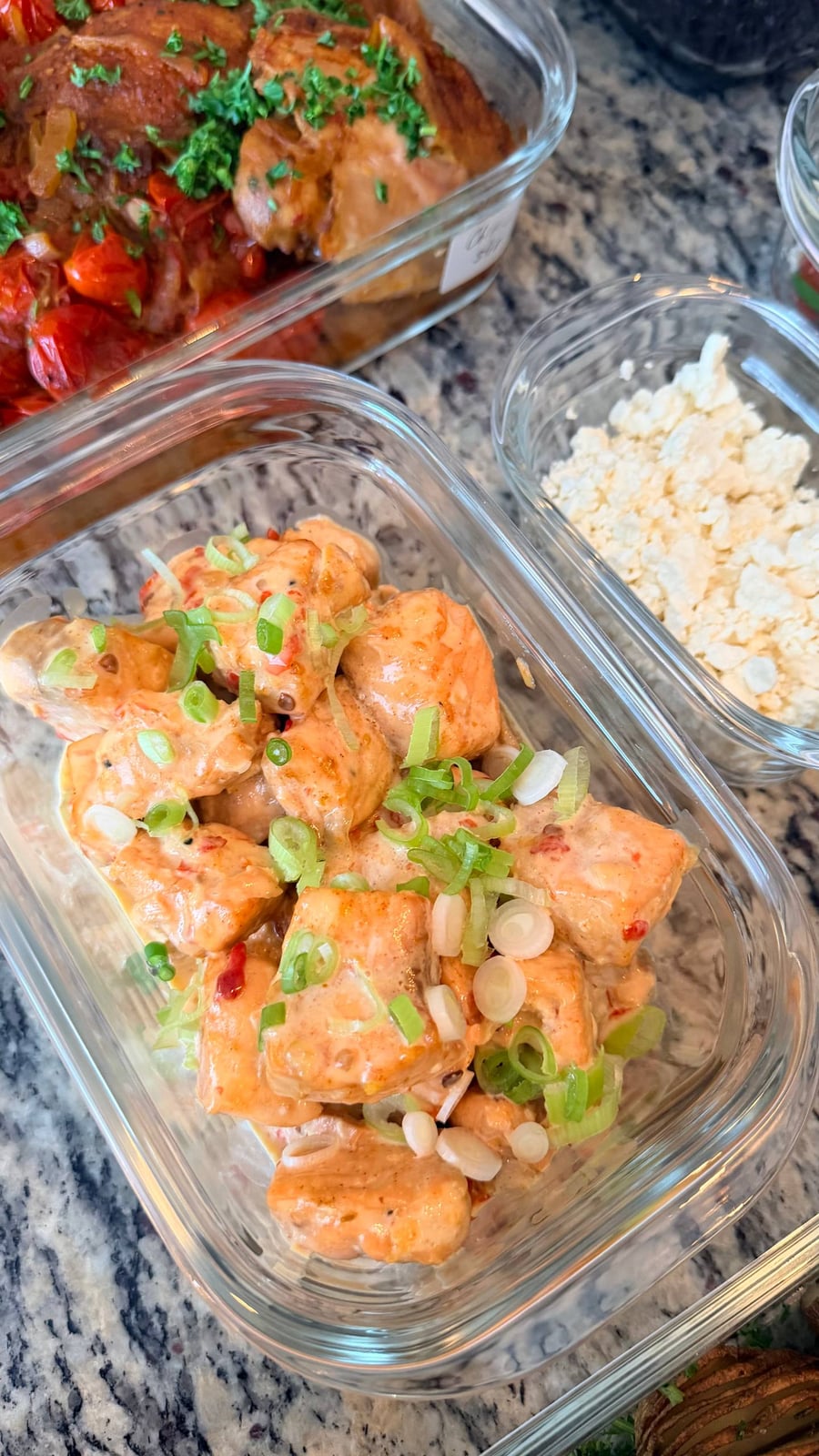 Glass meal prep containers filled with garlic salmon bites in bang bang sauce, topped with fresh scallions and accompanied by a side of crumbled feta.