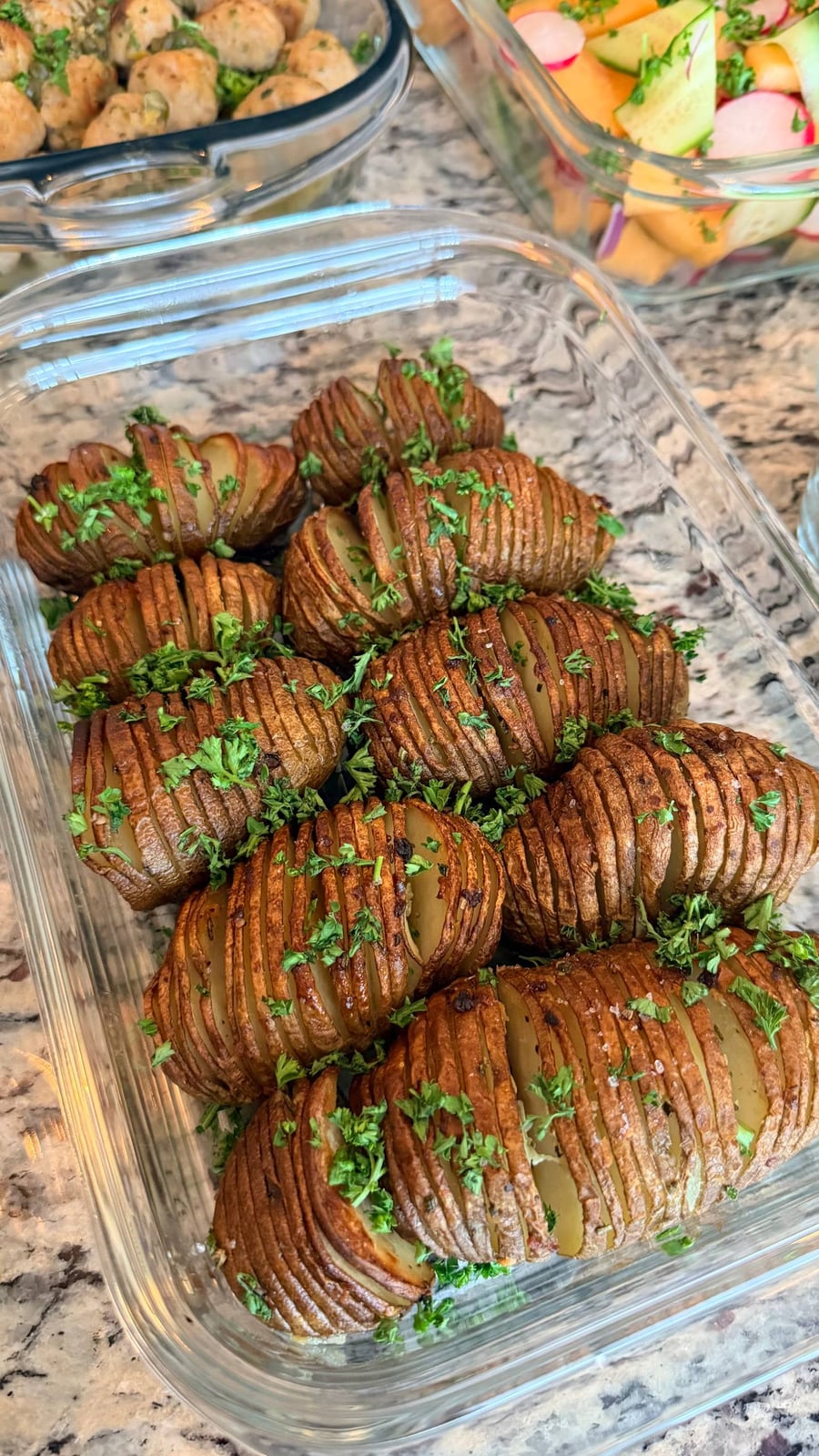A glass container filled with golden, crispy hasselback potatoes seasoned with garlic, rosemary, and fresh green parsley herbs for a delicious presentation.