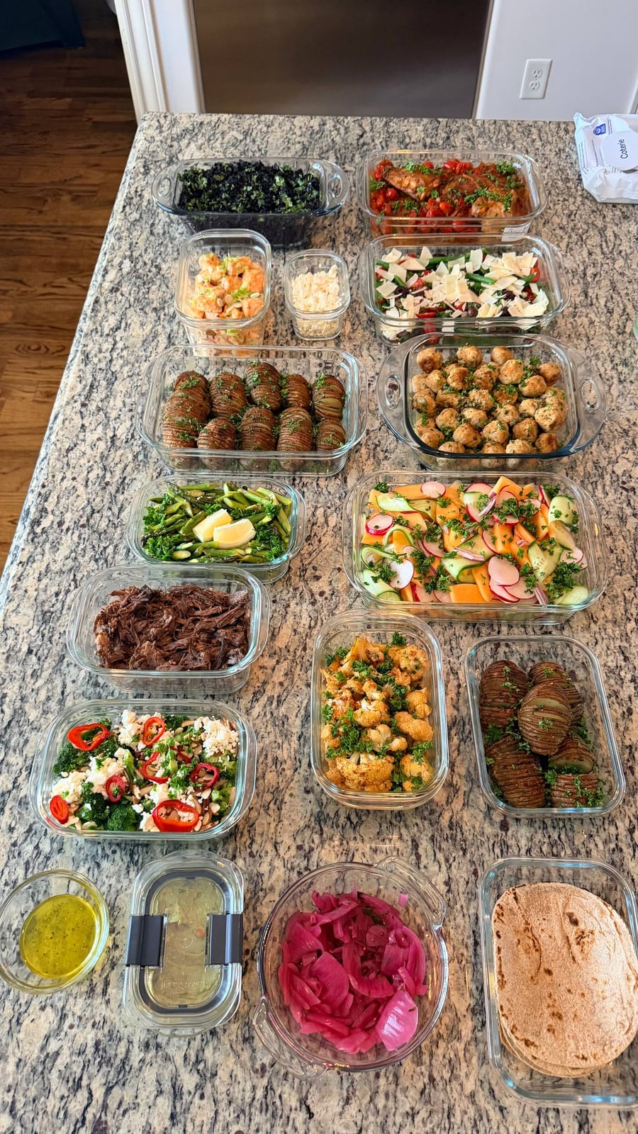 A variety of glass containers filled with chicken picatta meatballs, garlic salmon, shredded beef, roasted potatoes, fresh salad, and roasted vegetables on a granite countertop.