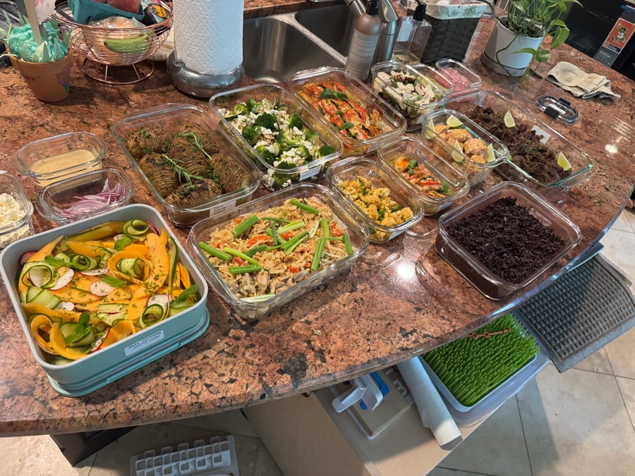 A variety of glass meal prep containers filled with garlic salmon bites, chicken pad thai, shredded beef, roasted potatoes, and a fresh melon salad.