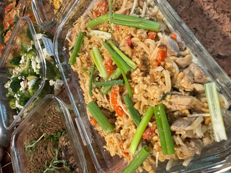 A glass container features chicken peanut Pad Thai noodles topped with crunchy crushed peanuts, vibrant sliced bell peppers, and fresh chopped green onions.