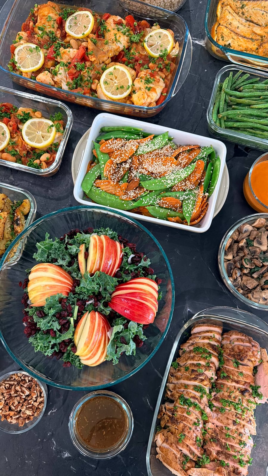 A variety of glass containers featuring harissa white fish with chickpeas, orange barramundi, sliced herb-crusted pork tenderloin, carrot stir-fry, and an apple kale salad.
