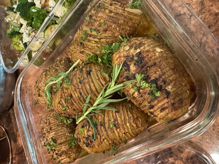A glass container filled with golden, crispy hasselback potatoes seasoned with roasted garlic and fresh rosemary sprigs, alongside a side of sautéed broccoli and zucchini.