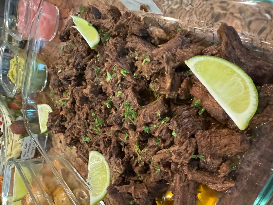 A large glass dish filled with tender Mexican style shredded beef garnished with fresh lime wedges and a sprinkling of chopped green herbs.