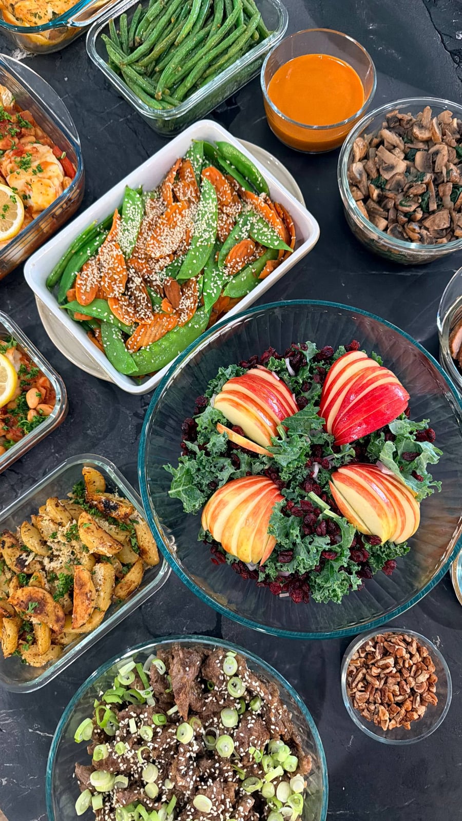 A variety of meal prep dishes including teriyaki beef, kale apple cranberry salad, sesame carrot snow peas, and garlic herb potato wedges.