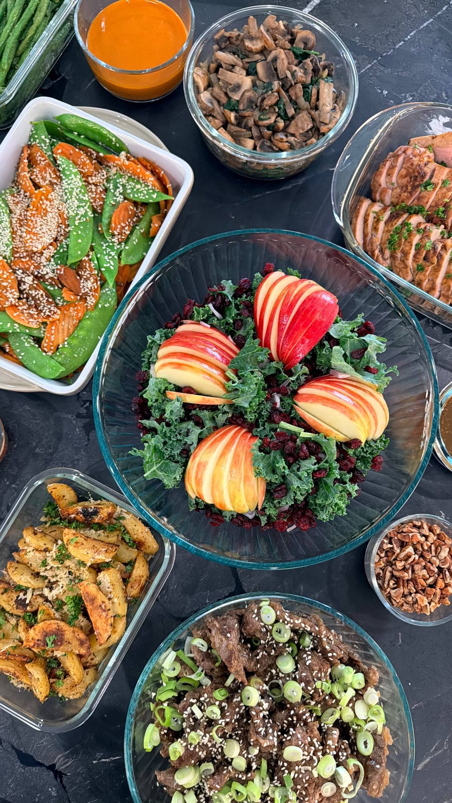 A spread of meal-prep dishes including teriyaki beef with scallions, a vibrant kale salad with sliced apples, baked pork tenderloin, and roasted garlic potato wedges.