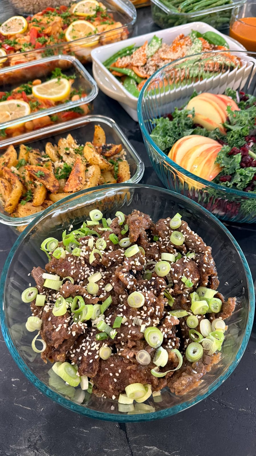 A glass bowl filled with tender teriyaki beef sirloin strips, garnished with fresh sliced scallions and toasted sesame seeds, served alongside varied meal sides.