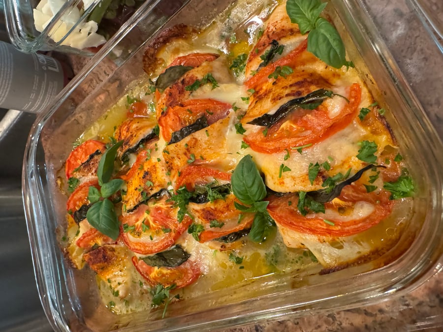 A glass baking dish containing tomato basil mozzarella chicken breasts, topped with fresh green basil leaves and herbs, served in a clear rectangular container.