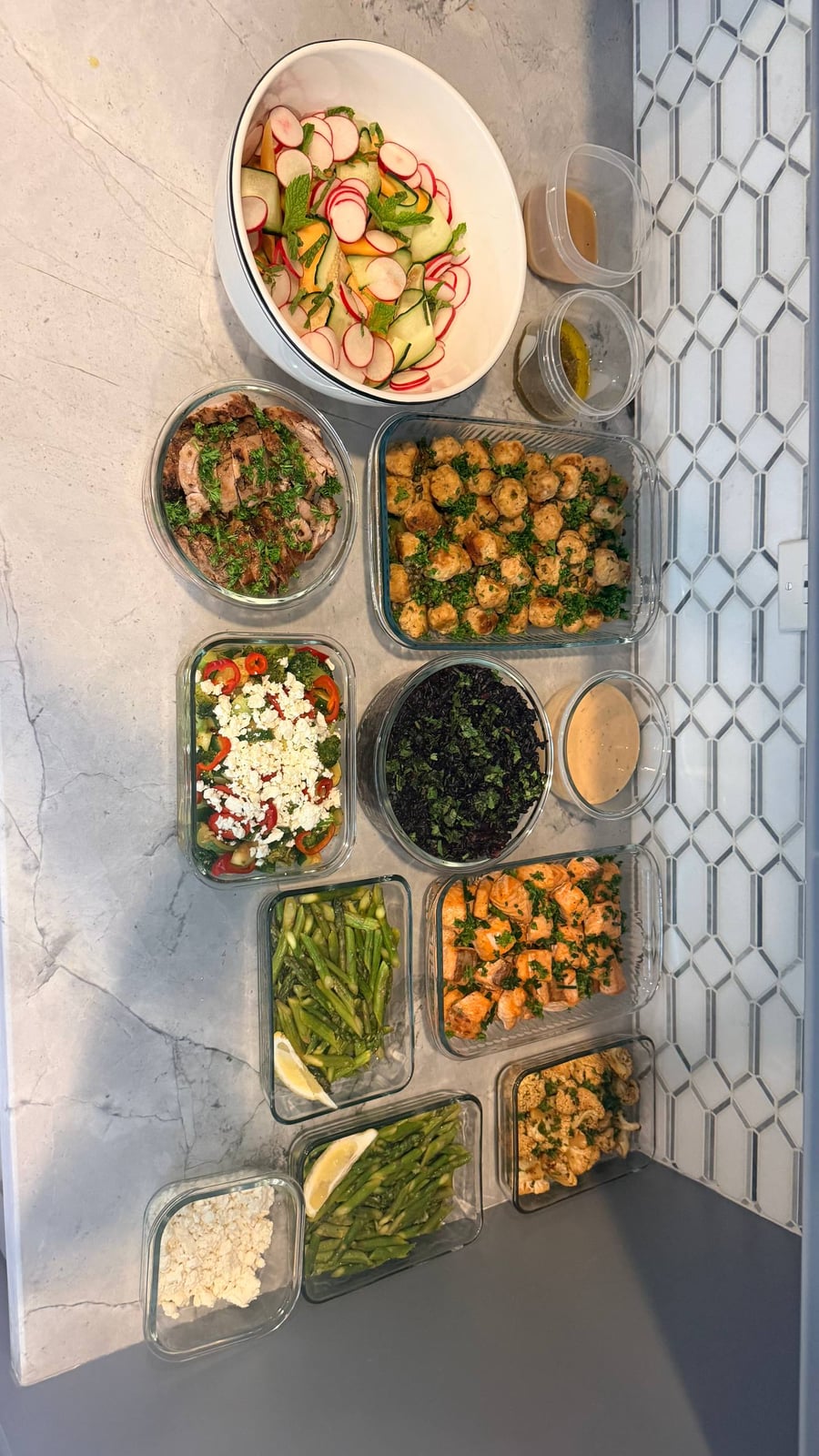 A variety of prepared dishes including chicken meatballs, garlic salmon, pork tenderloin, black rice, and a colorful salad served in glass containers.