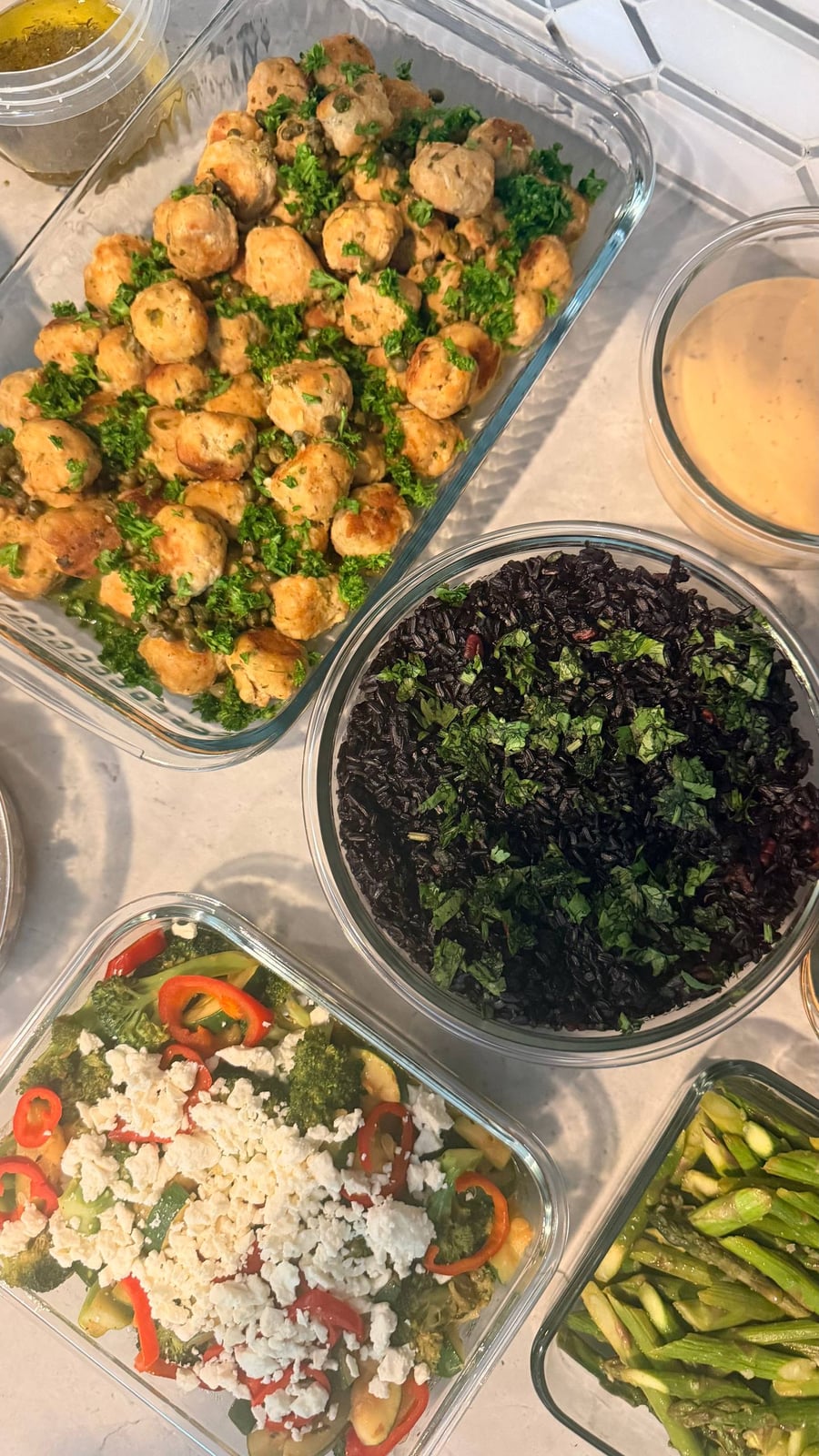 Glass containers featuring chicken picatta meatballs with capers, cilantro almond black rice, chili seared broccoli and zucchini, and ginger garlic asparagus stir-fry.