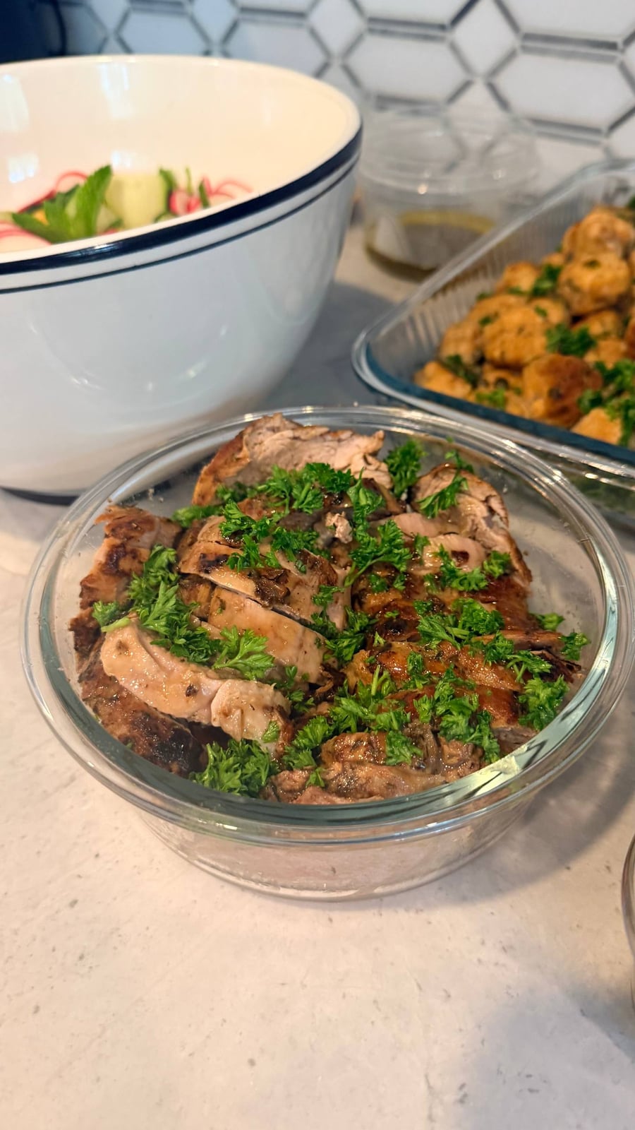 A glass bowl containing sliced pork tenderloin with maple mustard sauce, garnished with fresh parsley, alongside a bowl of melon salad and salmon bites.