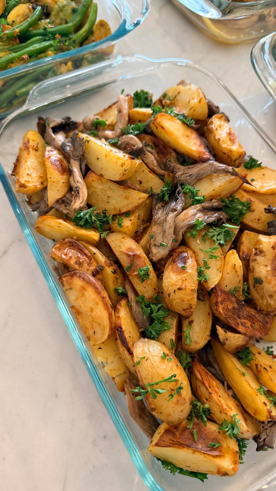 A glass container features golden-brown baked fingerling potatoes and savory oyster mushrooms, garnished with fresh green parsley on top of the roasted vegetables.