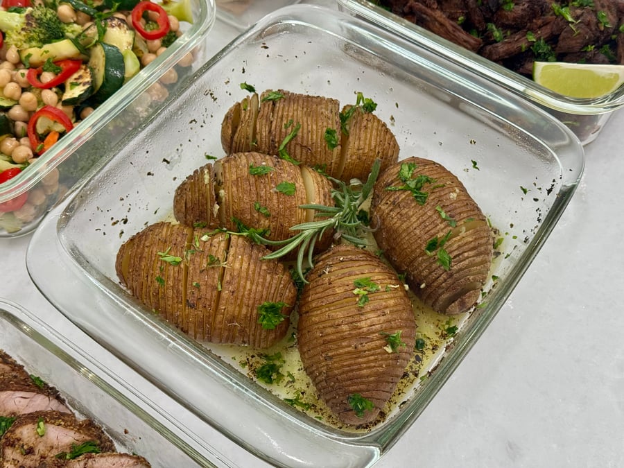 Four golden-brown hasselback potatoes seasoned with fresh rosemary and parsley, served in a glass container.