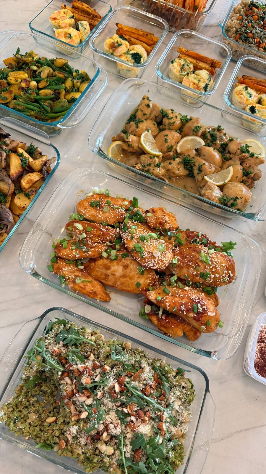 An overhead view of multiple glass containers filled with honey garlic chicken, saffron chicken, arugula pesto farro, and assorted vegetable sides on a counter.