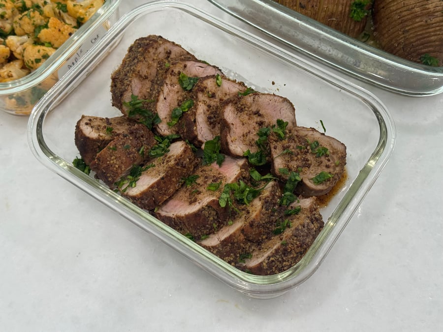 A glass container holding tender slices of pork tenderloin coated in a maple mustard crust, garnished with fresh herbs.