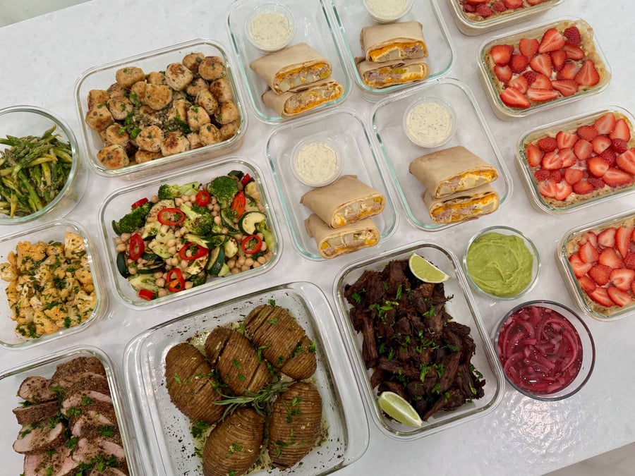 A variety of glass meal prep containers featuring seasoned proteins, side dishes, breakfast burritos, and strawberry baked oats arranged on a white counter.