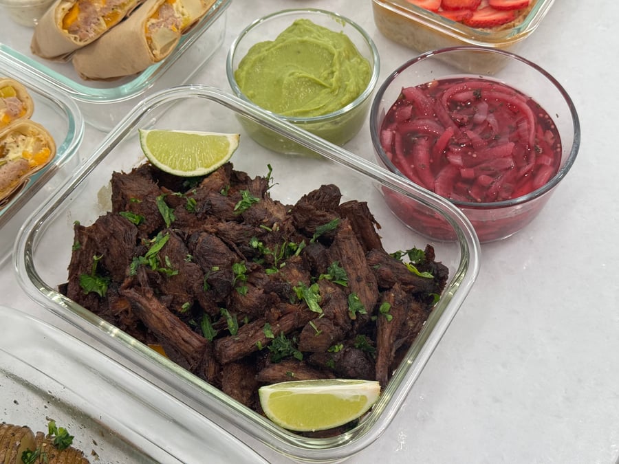 A glass container of tender shredded beef garnished with fresh lime wedges, served alongside small bowls of guacamole and pickled onions.
