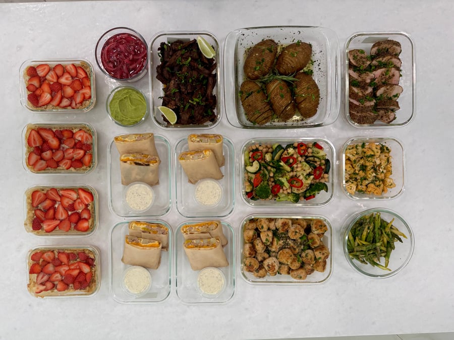 A variety of glass containers holding breakfast, protein, and side dishes including strawberry baked oats, shredded beef, meatballs, and roasted vegetables set on marble.