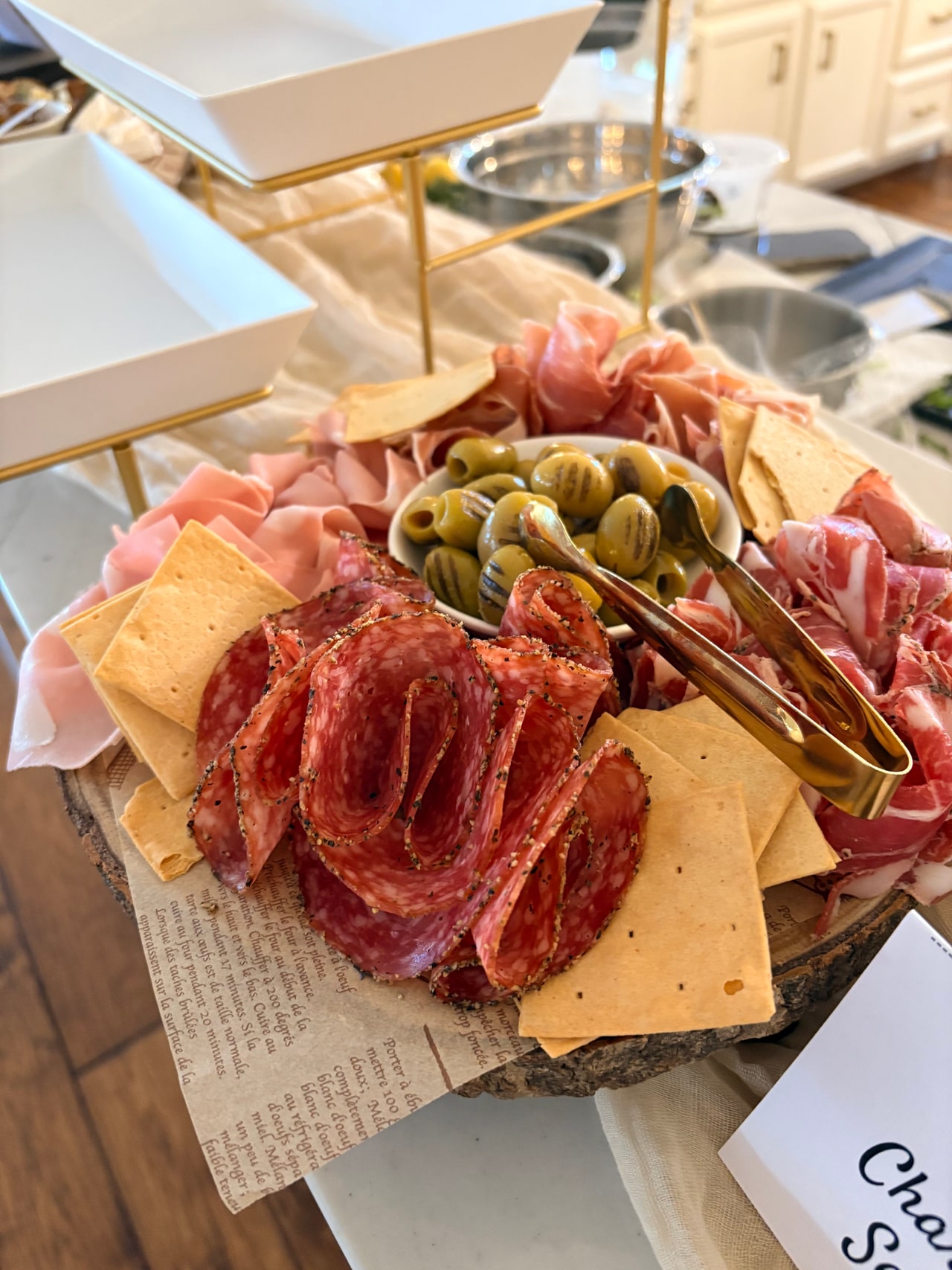 A wooden charcuterie board with cured meats, cheeses, olives and crackers beside the appetizer stands.
