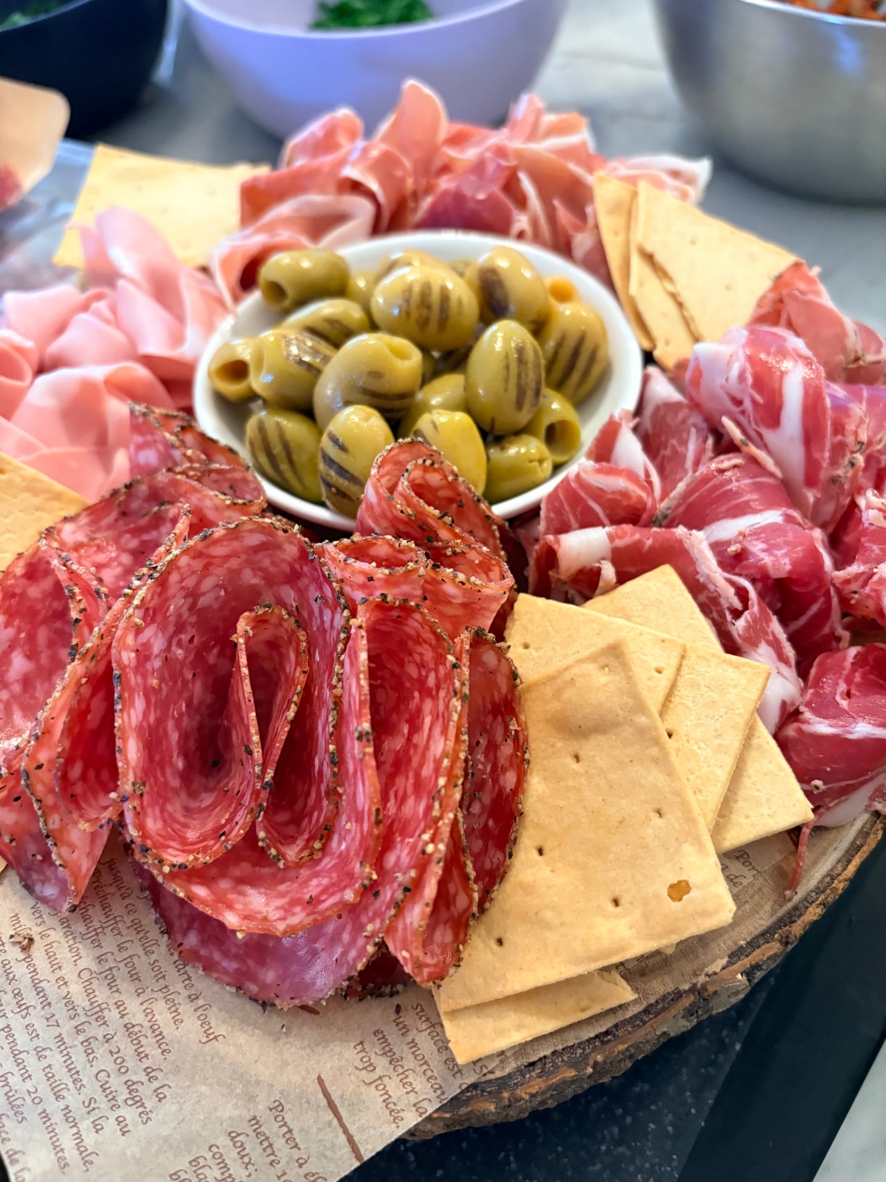 A round wooden board of sliced cured meats, cheeses, olives, crackers and a bowl of preserves.
