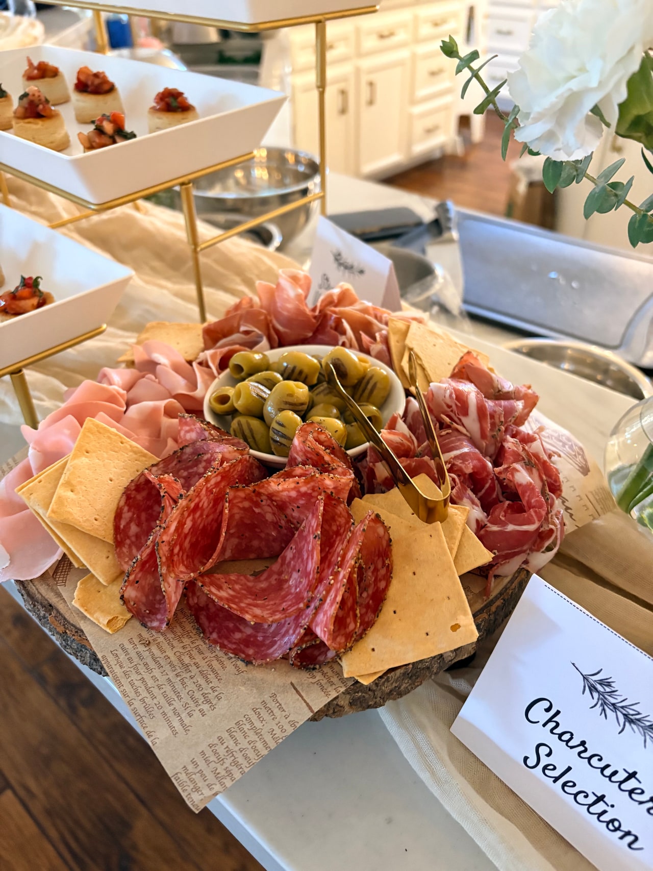 Cheese and cured meats on a round board in front of tiered strawberry vol-au-vents and other appetizers.