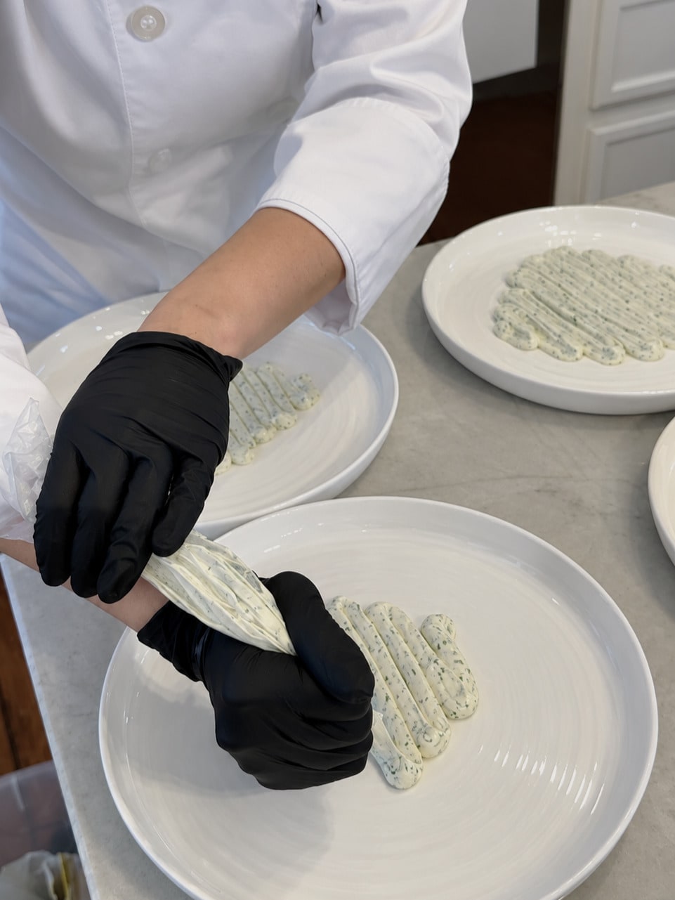 A chef wearing black gloves pipes a herb-speckled cream onto white dinner plates.