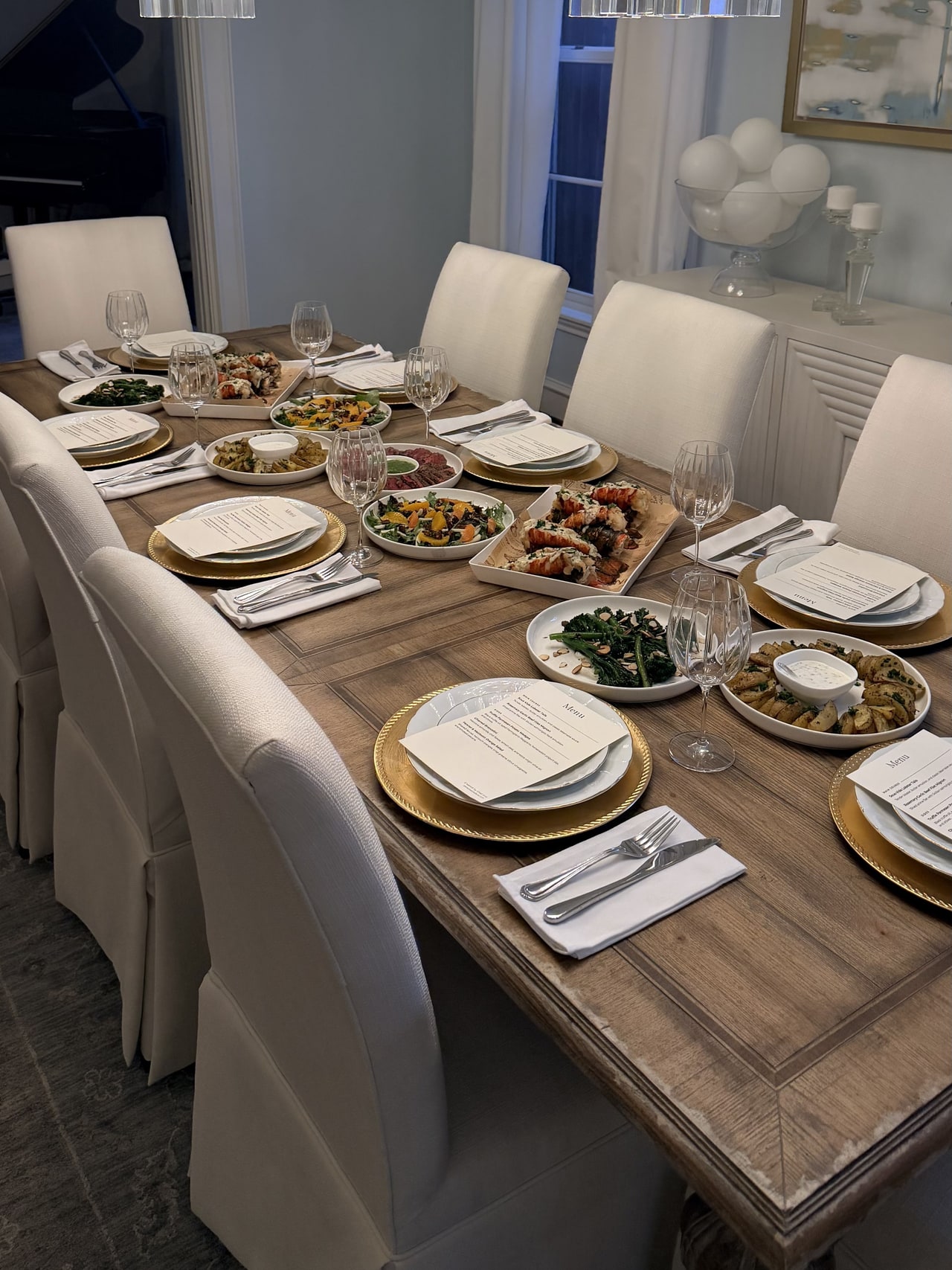 A home dining table set with printed menus, gold-rimmed plates, glassware and shared dinner platters.