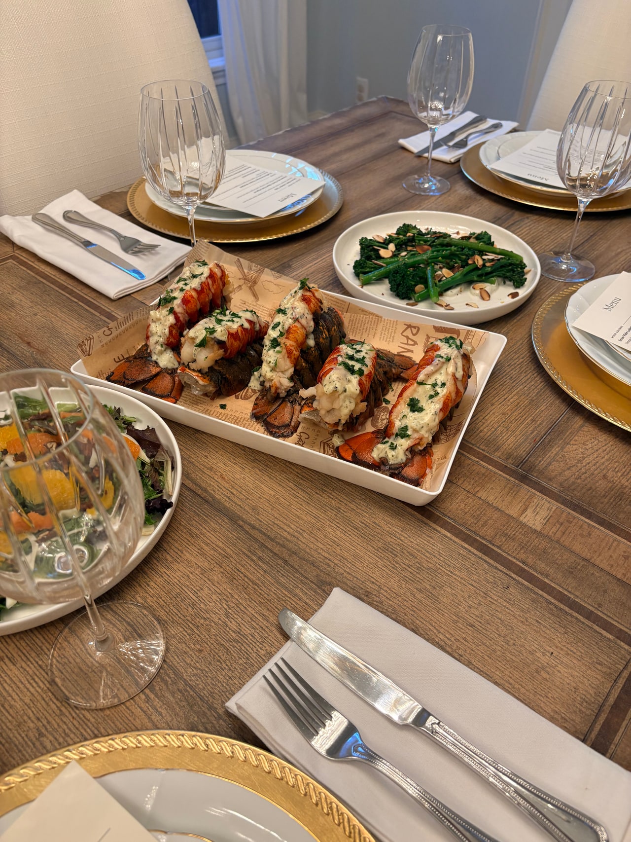 Lobster tails on a wooden board with broccolini, fennel and orange salad, and place settings around them.