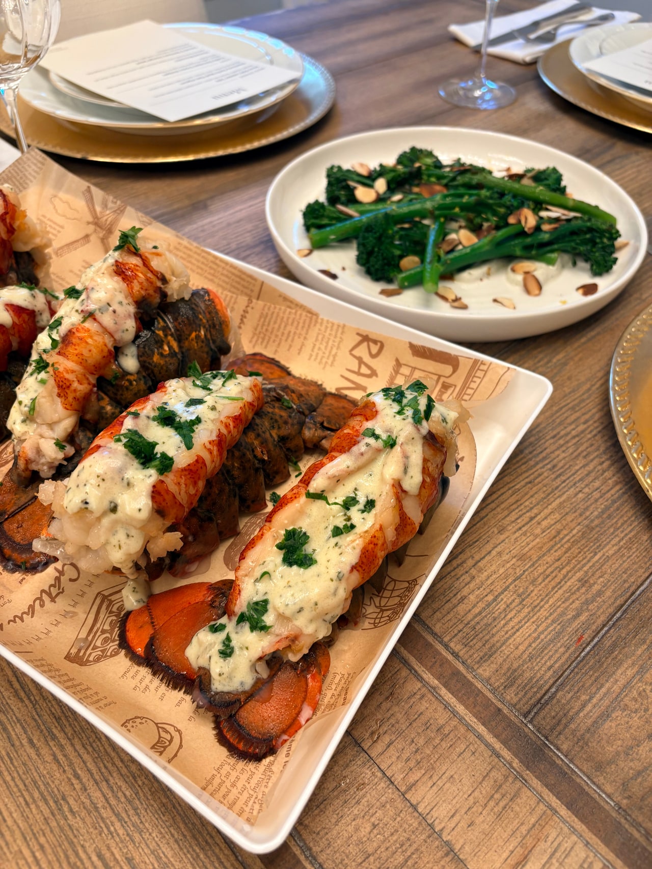 Sauced lobster tails on a wooden serving board beside a blue-and-white platter of broccolini.