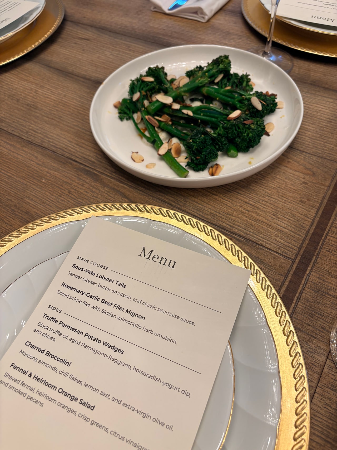 A printed surf and turf menu on a folded napkin and gold-rimmed plate, beside a platter of broccolini.