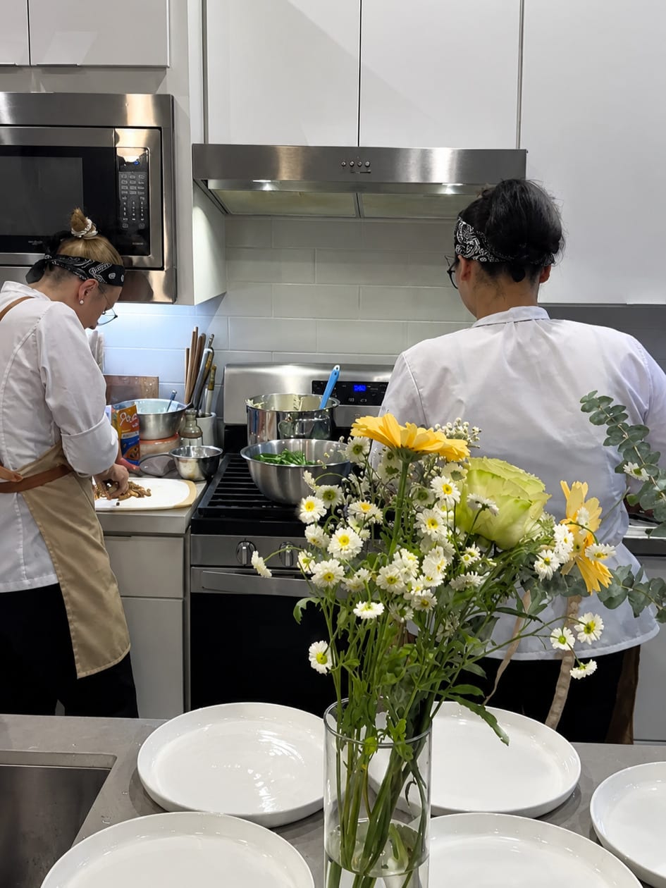 Two chefs work at the stove in a kitchen beside a vase of flowers.