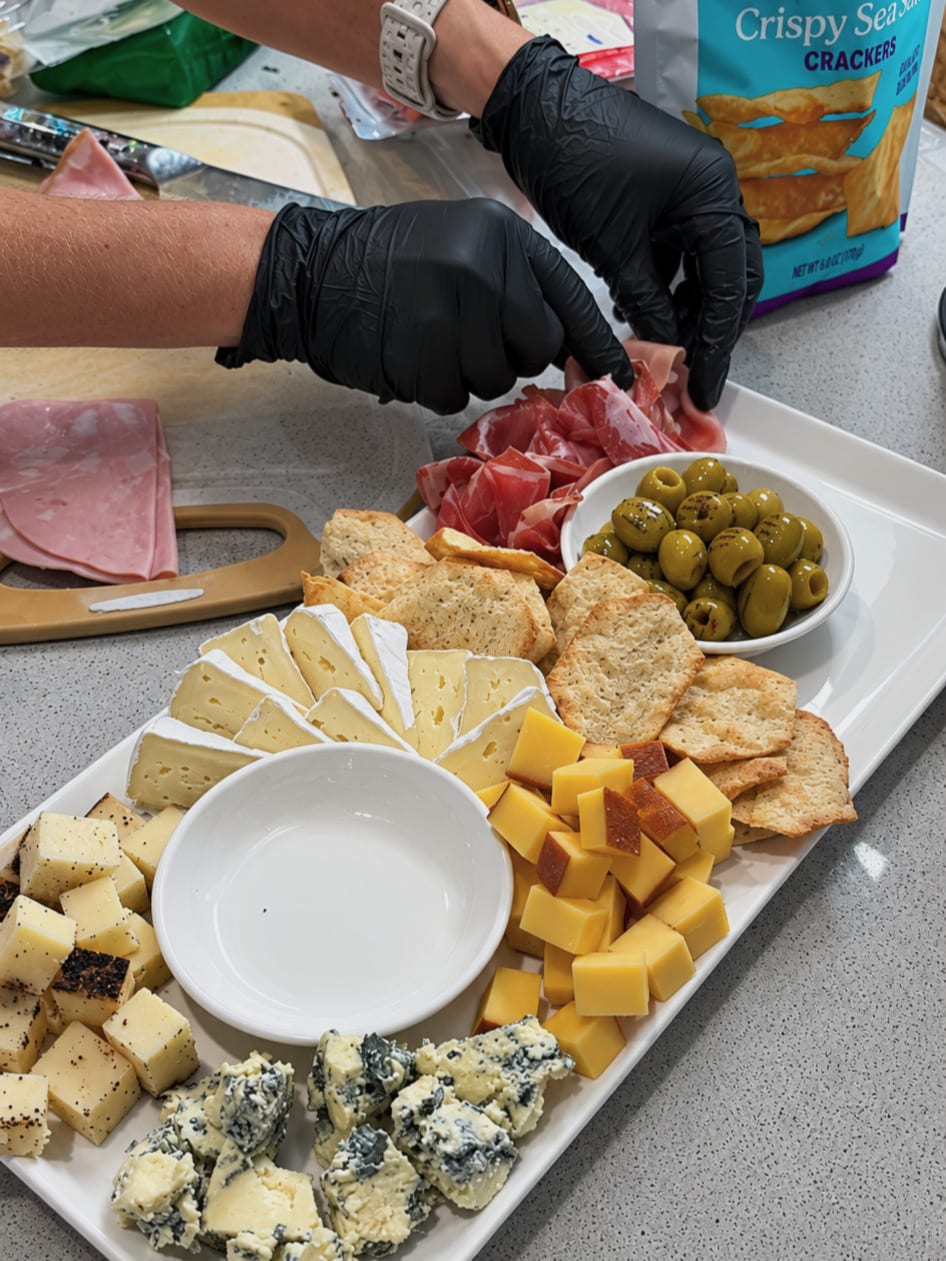 Gloved hands arrange cured meats beside olives, cheese and crackers.