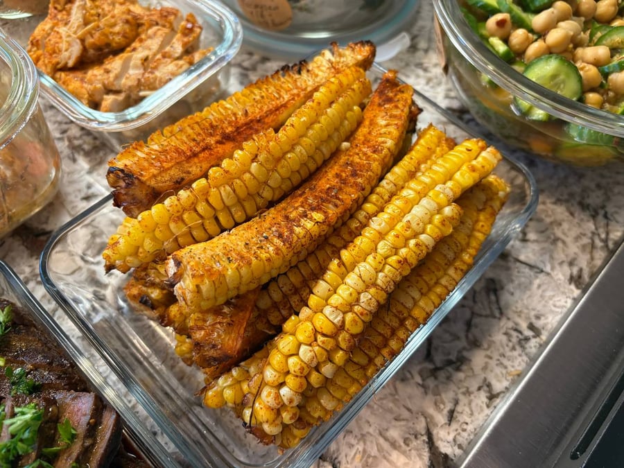 A glass container filled with paprika-seasoned corn ribs, alongside other containers holding sliced chicken, spicy cucumbers, and chickpeas.
