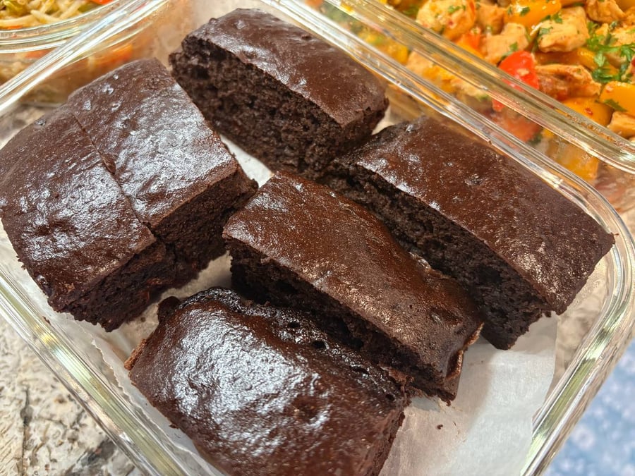 A glass container holds four thick squares of dark, moist-looking healthy protein brownies arranged neatly on parchment paper.