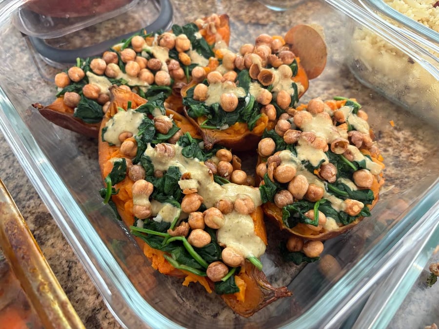 A glass container holds four roasted sweet potato halves stuffed generously with sautéed green spinach, protein-rich chickpeas, and a drizzle of savory tahini dressing.