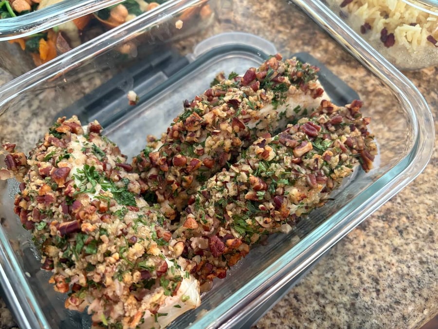 A glass meal prep container filled with two golden, pecan-crusted halibut fish fillets, seasoned with fresh herbs and chopped toasted nuts.