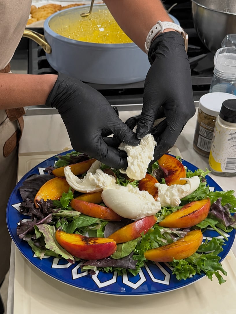 A chef wearing black gloves plates burrata over peaches and greens, with a dressing bowl nearby.