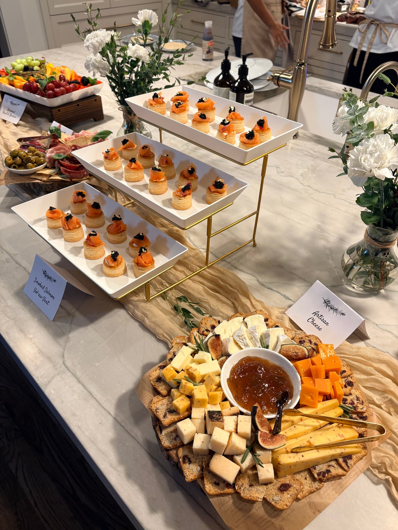 An appetizer display with tiered smoked salmon vol-au-vents, cheese, charcuterie and fruit on the kitchen counter.